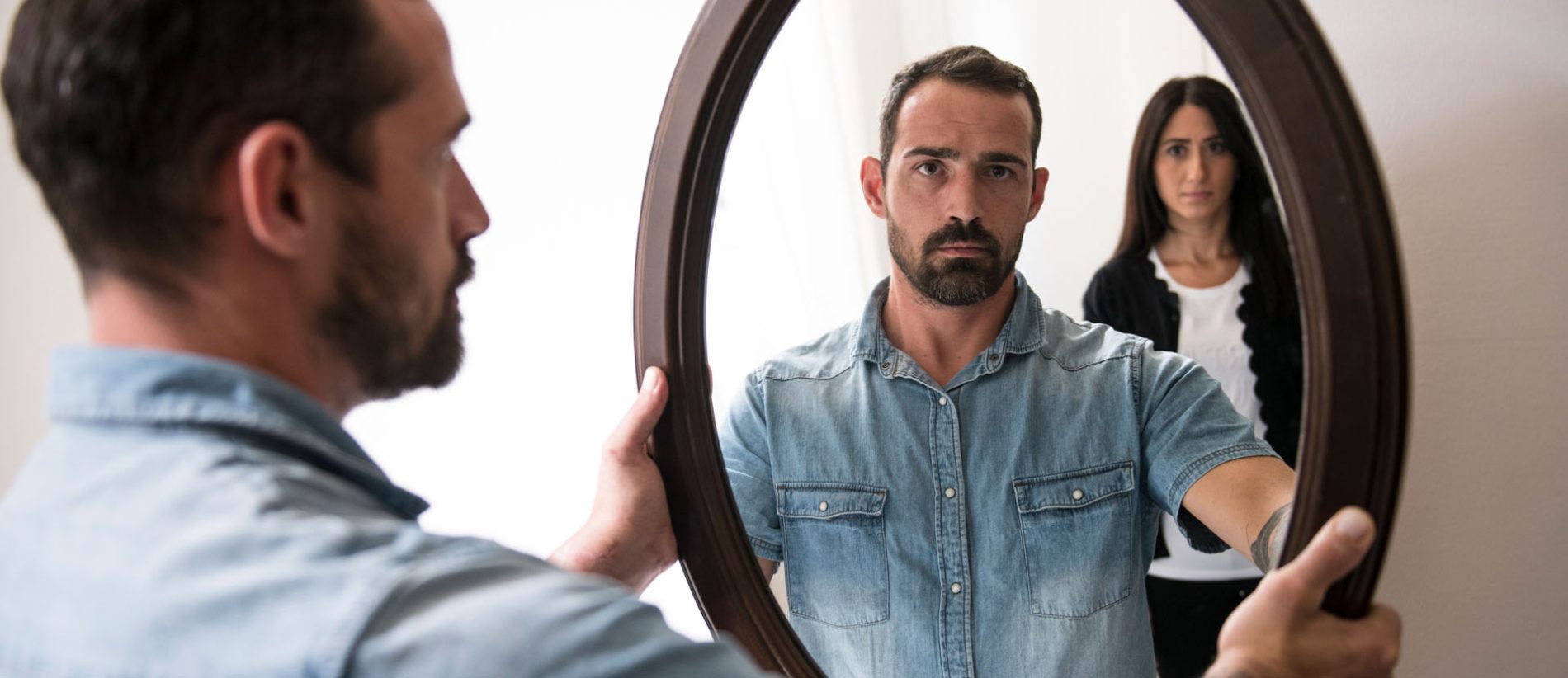 immagine di un uomo che si guarda allo specchio preoccupato e alle sue spalle una donna impaurita
